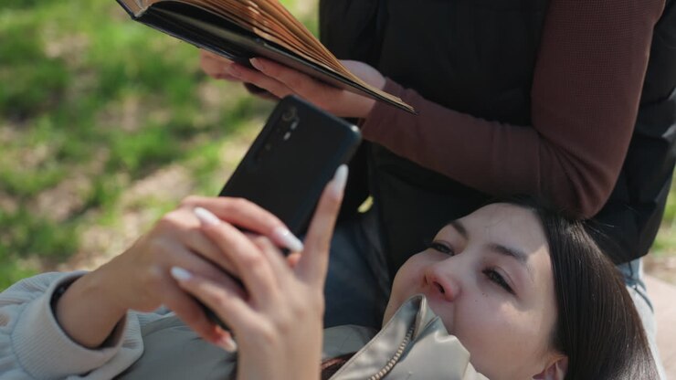 woman lying on bench scrolling smartphone with friend reading book, spring park grass and sunlight, casual jacket and touchscreen closeup, quiet study and leisure mood, candid restful moment