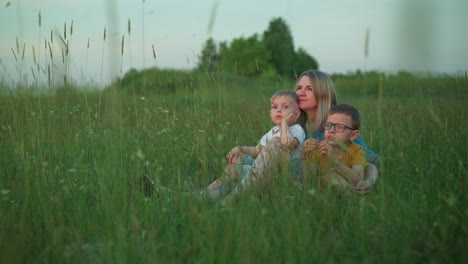 a mother in a blue gown sits in grass field with her two children. the older child, wearing a yellow top and glasses, and the younger child, dressed in a white top, are both seated closely to her