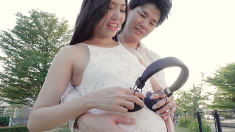 happy pregnant asian woman and husband with headphones on her belly in garden. 4k