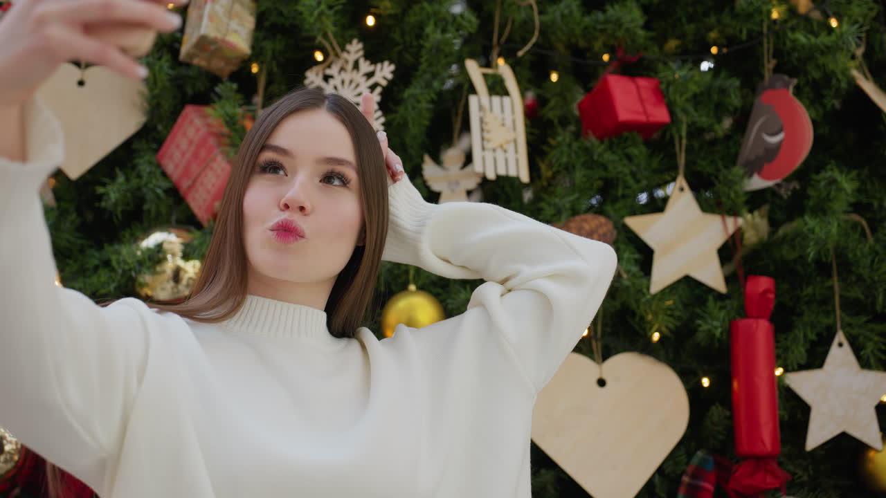 Stylish woman in white top adjusting her phone while taking a playful selfie in front of beautifully decorated Christmas tree with festive ornaments, striking a fun pose with hand on head