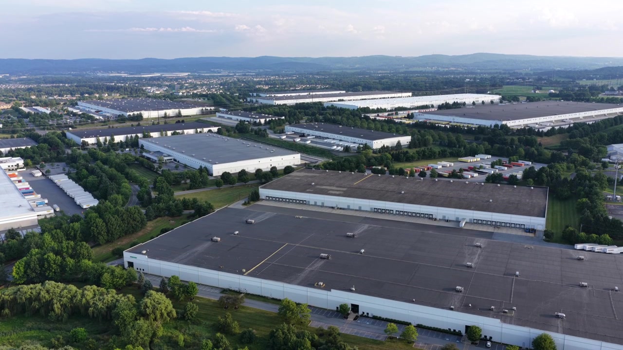 Rækker af lagerbygninger med fladt tag fylder industriparken, omkranset af træer i Allentown, Pennsylvania. Fjerne bjerge rejser sig bag spredte parkeringspladser og adgangsveje. Vidvinkelbillede fra stigende drone.