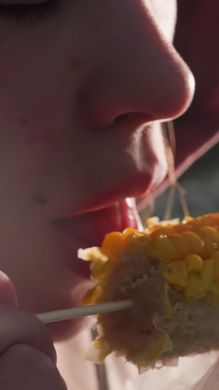 primer plano de una mujer comiendo maíz sostenido por un palo envuelto en plástico blanco con luces bokeh en un fondo borroso, vapor que se eleva desde el maíz recién cocido en un entorno al aire libre en un día soleado