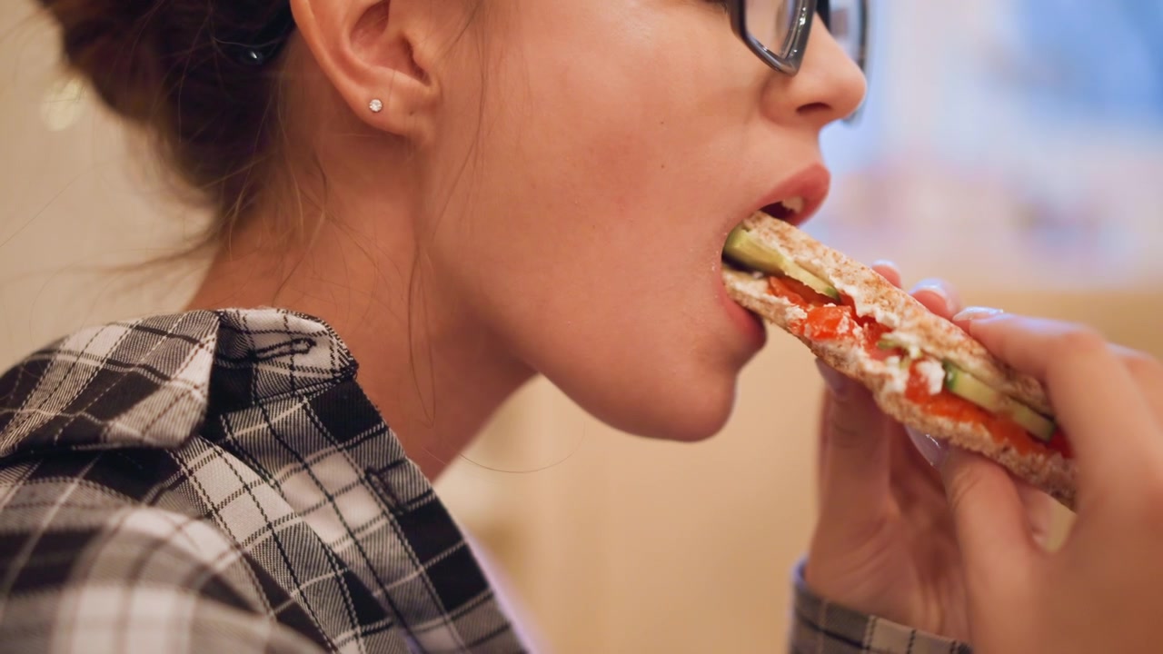Close up of woman eating food while wearing glasses and plaid shirt. Sandwich with visible ingredients like cucumber tomato is held in hand as woman takes bites. Soft indoor lighting creates