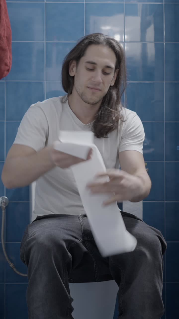 A man using toilet paper while sitting on a toilet in a blue-tiled bathroom