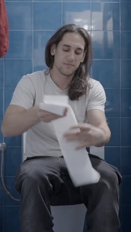 a man using toilet paper while sitting on a toilet in a blue-tiled bathroom