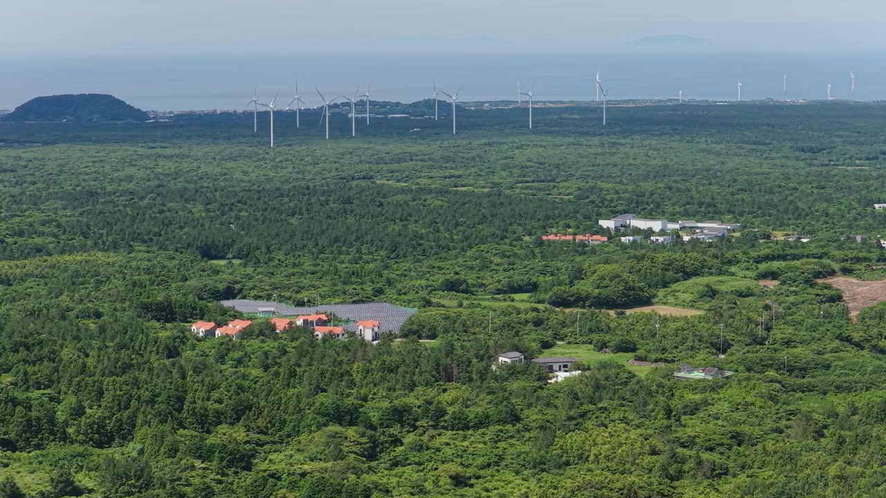 Horizontal elevated view of Dongbaek Dongsan Wetland in eastern Jeju Island, South Korea, showing dense forest, wind turbines, fields, buildings and the distant sea as the camera slowly trucks left