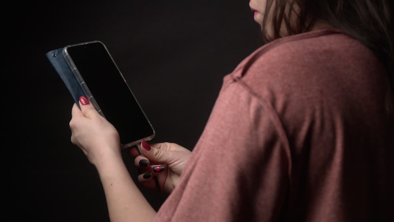 A woman in a cozy setting holds a smartphone, plugging in a charger. The dim lighting and focus on her hands and device create an intimate, detailed atmosphere
