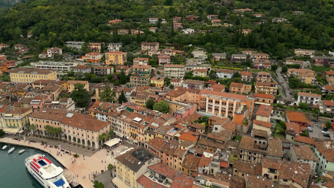 Vista aerea su una cittadina lacustre con pontile per barche, edifici storici e colline verdi.