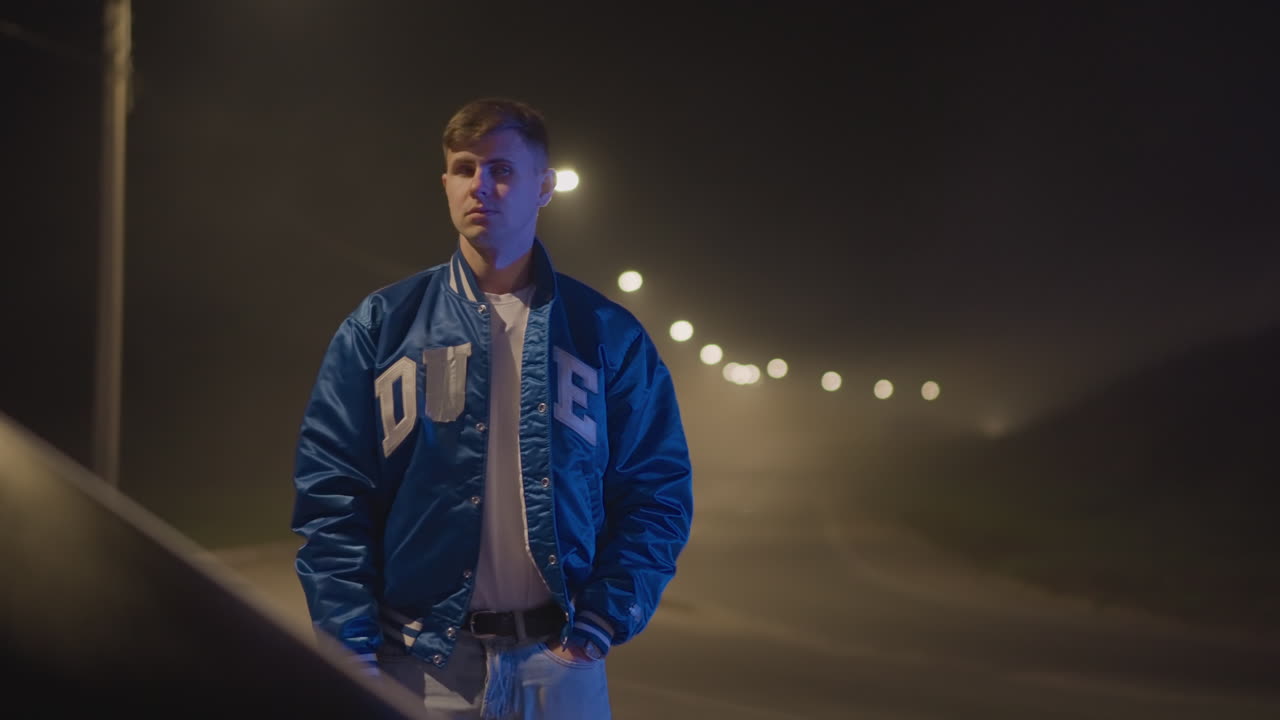 Bredt nattbilde av en hvit mann som står ved en tom vei, blå jakke som fanger gløden fra gatelykten, fjern bokeh-streng av lamper, kontemplativ holdning og sigarettliknende damp, filmatisk ensomhet sent på kvelden