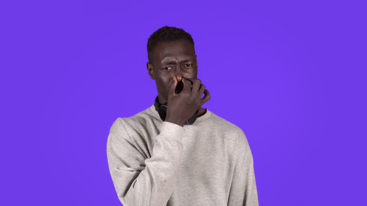 Afro american man isolated against blue background with headphones on neck smelling something stinky and disgusting, holding breath with fingers on nose. Bad smells concept