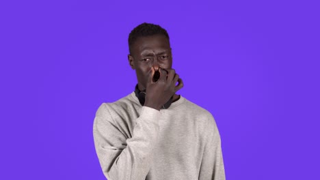 afro american man isolated against blue background with headphones on neck smelling something stinky and disgusting, holding breath with fingers on nose. bad smells concept