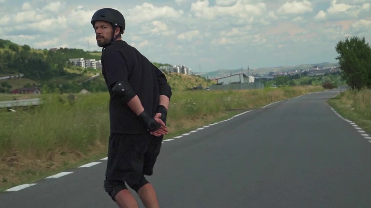 A skateboarder moves smoothly down a winding road, surrounded by open fields and distant hills.