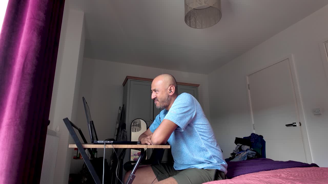A man sitting in a bedroom working remotely on a laptop at a desk.
