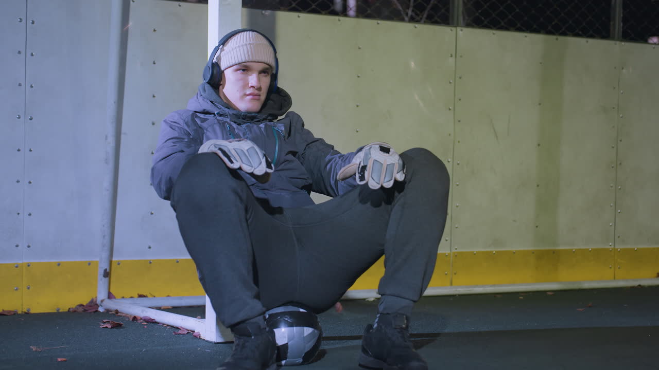 Young man resting against goalpost while listening to music with headphones, shaking head in rhythm, wearing hoodie and gloves during night session, soccer ball beneath