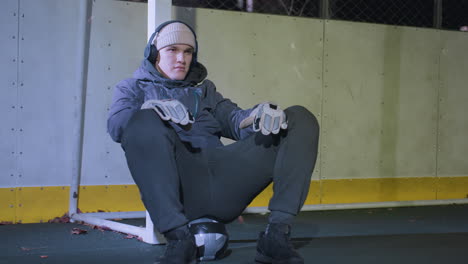 joven descansando contra el poste de la portería mientras escucha música con auriculares, sacudiendo la cabeza en el ritmo, con capucha y guantes durante la sesión nocturna, bola de fútbol debajo