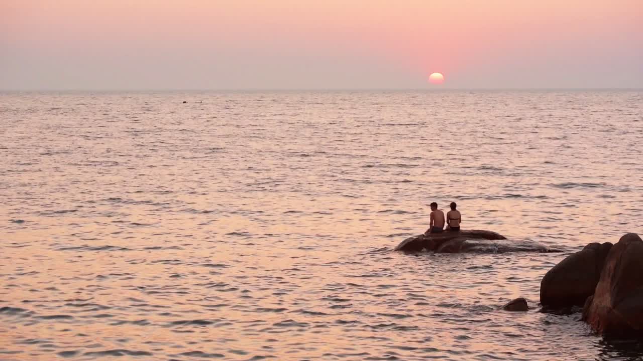 A Couple Sitting In The Sea Rock Watching The Sunset And The Arabian Waters In Goa, India. -wide shot