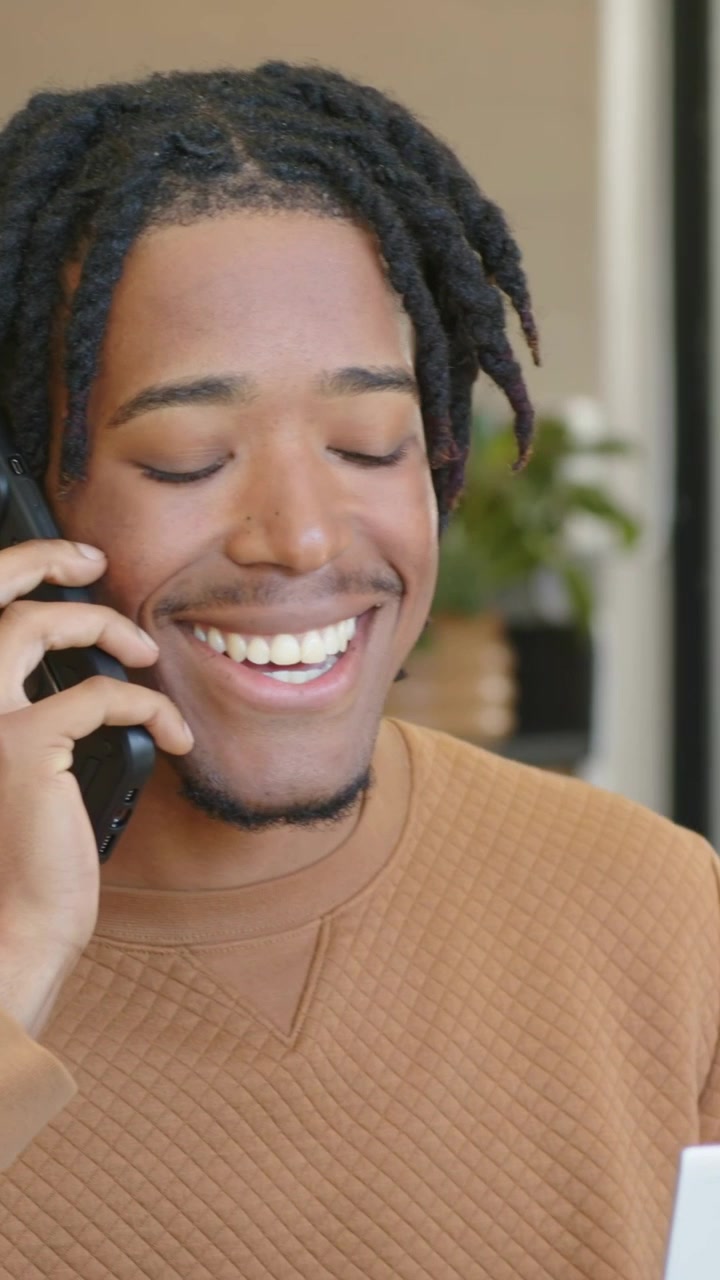 Vertical video: Reading man in tan sweater with locs testing mic on throat, calling cell in kitchen