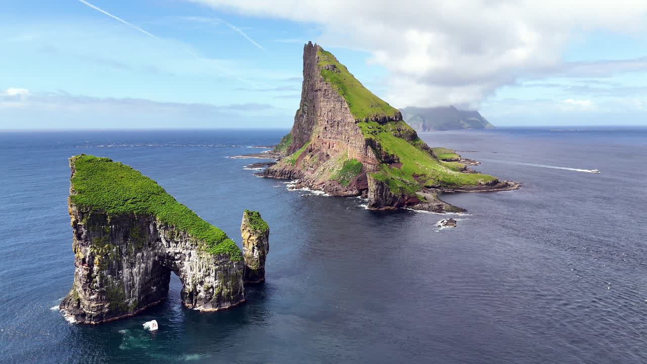 Cinematic aerial view of Drangarnir sea stacks rising dramatically from the Atlantic Ocean near Vágar, Faroe Islands, showcasing rugged cliffs, lush green slopes, and misty Nordic seascape