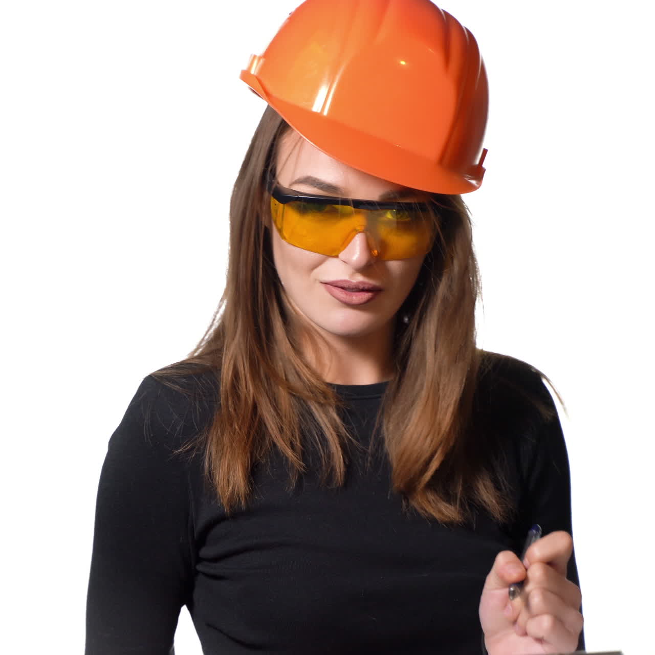 An entertaining woman in orange glasses and hard hat is holding a folder and pen in her hands and talking with showing something on a white studio background.