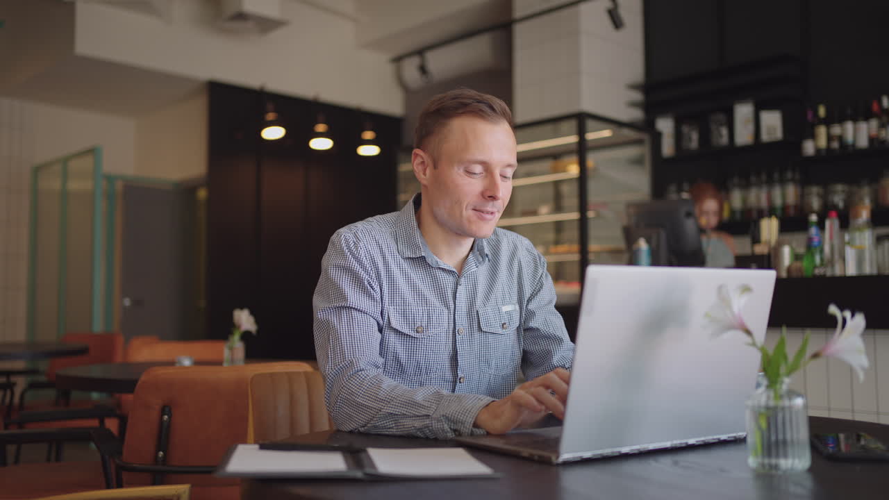 A young man in a shirt is sitting at a table with a laptop and typing on the keyboard. A student can study remotely. A businessman conducts his business remotely
