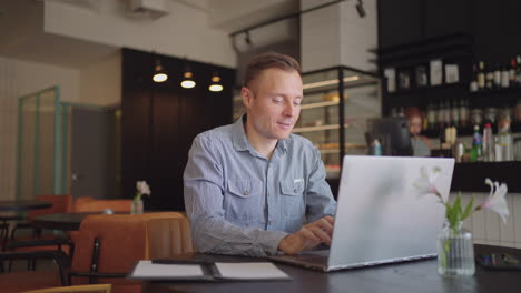 a young man in a shirt is sitting at a table with a laptop and typing on the keyboard. a student can study remotely. a businessman conducts his business remotely