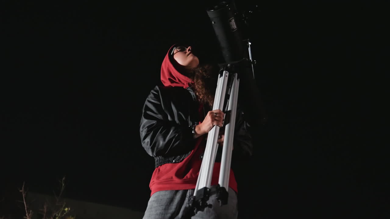 observer with telescope on city night, searching stars from grassy lot beside illuminated apartment building while adjusting tripod and eyepiece, quiet curiosity and scientific wonder in lowlight