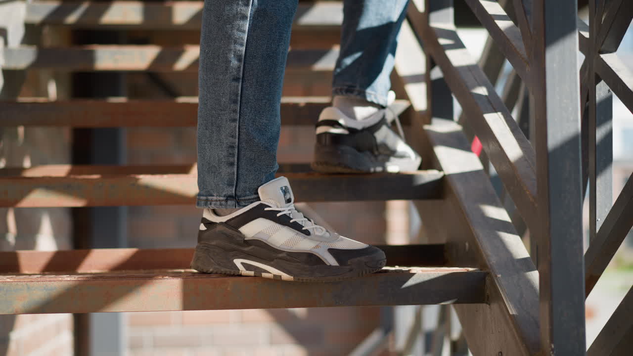 Legs in blue jeans and black white canvas sneakers standing on rusty metal staircase with geometric railing as sunlight casts sharp shadows creating contrast in urban outdoor scene