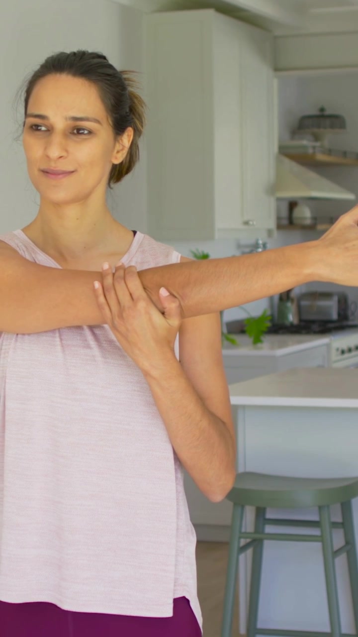 Video vertical: Mujer con camiseta sin mangas tirando, entrelazando y empujando los brazos en la cocina para calentar los hombros