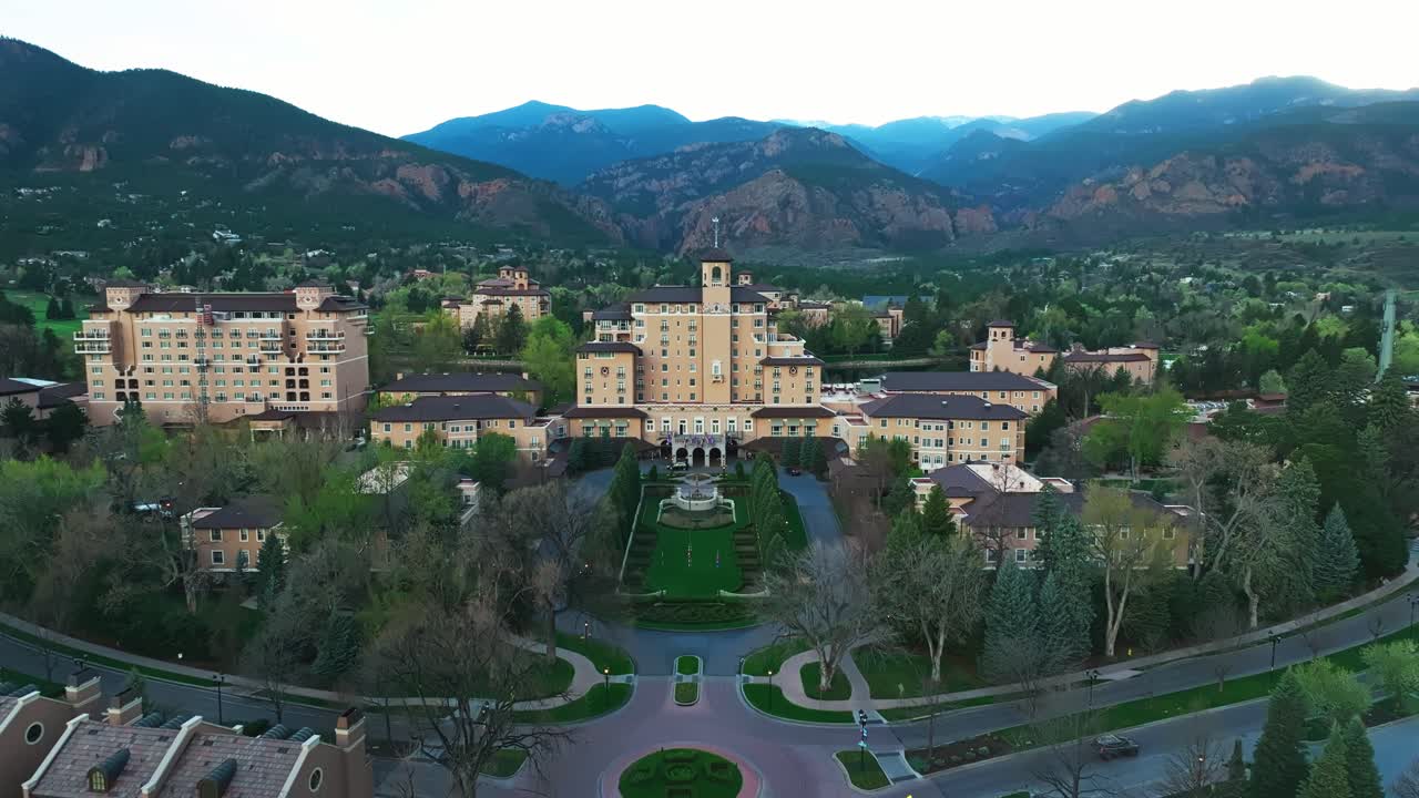 Sunlit Broadmoor estate nestled in wooded landscape with calm water beside it, aerial approach