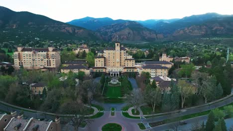 sunlit broadmoor estate nestled in wooded landscape with calm water beside it, aerial approach
