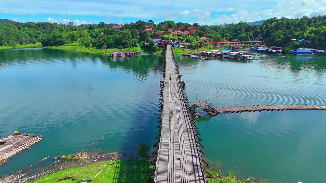 Static drone shot of Mon Bridge in Sangkhlaburi, Thailand, with people walking across the iconic wooden span above the river, surrounded by lush green hills and village houses