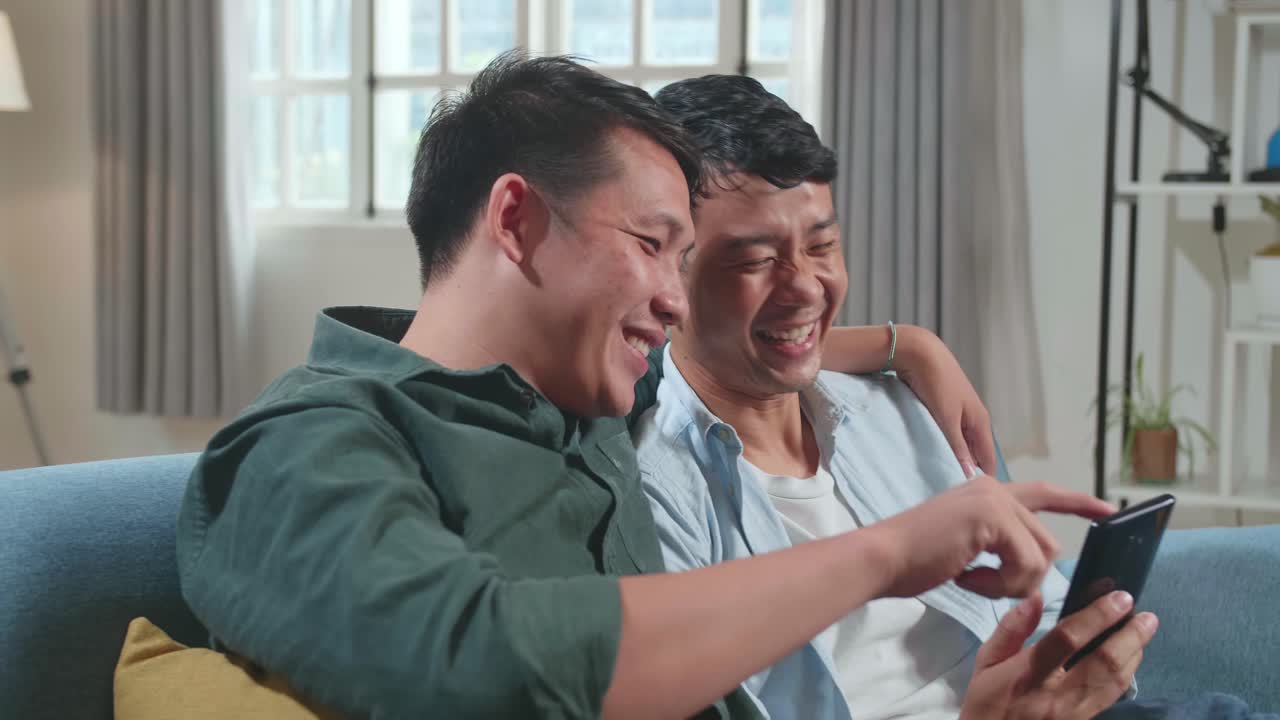 Portrait Of Asian Gentle Gay Couple Using Mobile Phone, While Sitting On A Couch In Cozy Stylish Apartment. Adult Boyfriends Online Shopping On Internet, Watching Funny Videos On Streaming Service