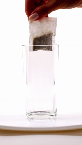 mano sumergiendo una bolsa de té en un vaso claro de agua en un plato blanco, fondo blanco