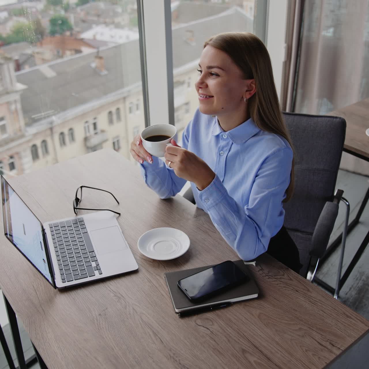 Donna bionda seduta alla scrivania in ufficio che tiene in mano una tazza di caffè. Signora calma e rilassata che si gode la pausa pranzo sul posto di lavoro. Vista dall'alto