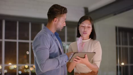 Focused-diverse-male-and-female-colleague-standing-talking-and-using-tablet-at-night-in-office