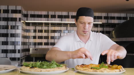 Front-View-Of-A-Chef-Placing-Basil-Leaves-On-Baked-Pizzas-And-Then-Looks-At-The-Camera