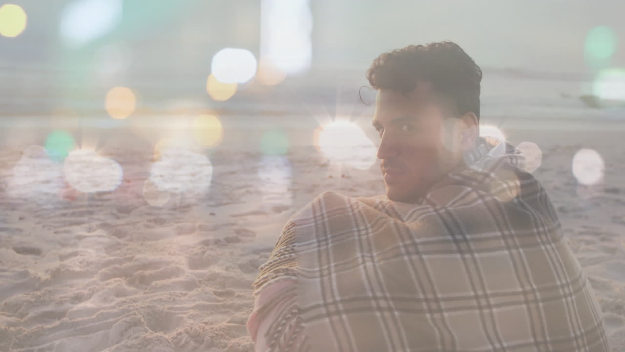 Sitting on beach, man wrapped in blanket with animation of bokeh lights