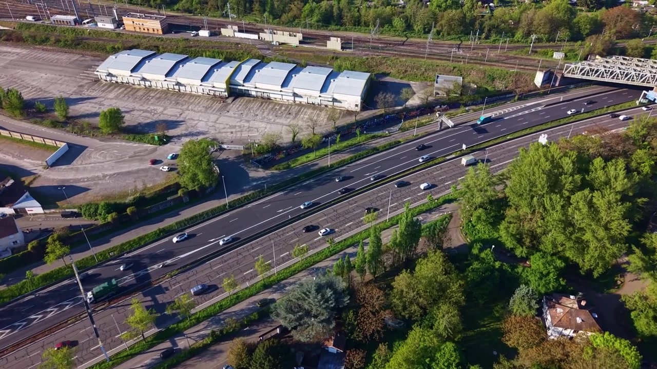 Drone flyover of cars and trucks move along the National 4 highway beside warehouses, railway lines and suburban houses, Strasbourg, France