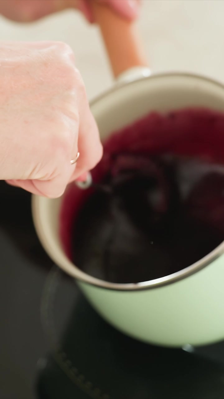 Primo piano di mano che mescola un liquido rosso scuro in una pentola verde chiaro. La miscela densa viene mescolata costantemente mostrando cambiamenti graduali. Lo sfondo rimane neutro e sfocato per tutto il tempo