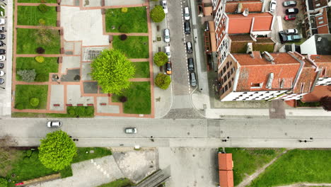 aerial view of a cityscape, featuring a crossroads with cars, a pedestrian area with benches and green trees, surrounding residential buildings, and distinct architectural elements