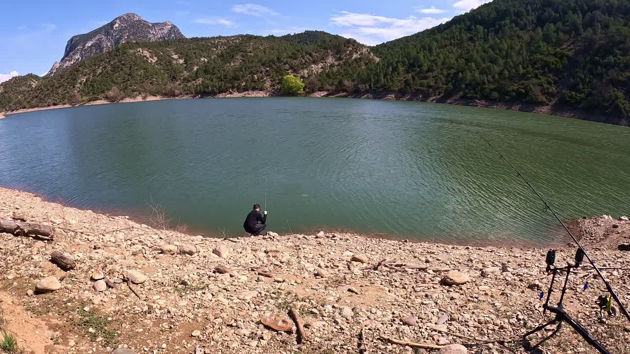 A lone angler stands on a rocky lakeshore casting a line into the calm, shimmering water of a mountain lake, surrounded by lush green forests and rugged hills on a clear day