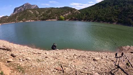 a lone angler stands on a rocky lakeshore casting a line into the calm, shimmering water of a mountain lake, surrounded by lush green forests and rugged hills on a clear day