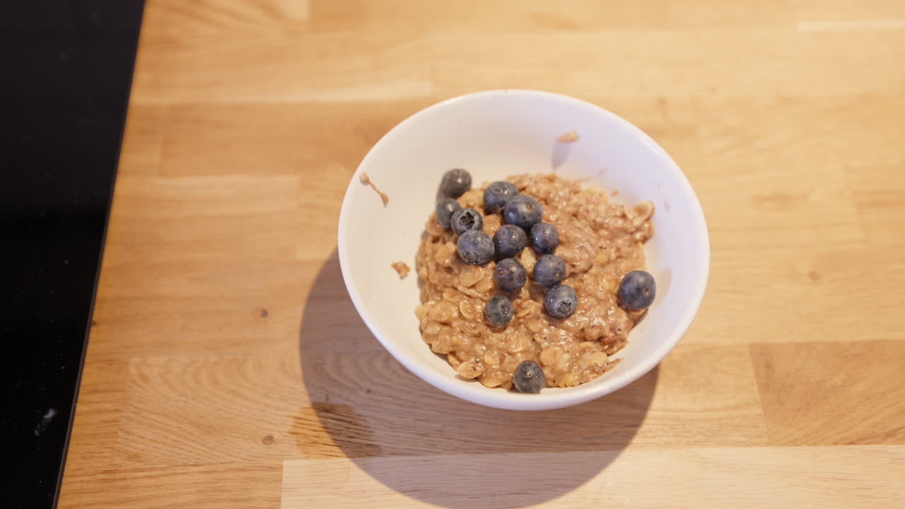 Mirtilli freschi aggiunti sopra il porridge di avena al cacao in una ciotola. Colazione fatta in casa sana e colorata. Pasto mattutino nutriente con guarnizione di frutta, filmato reale e autentico.