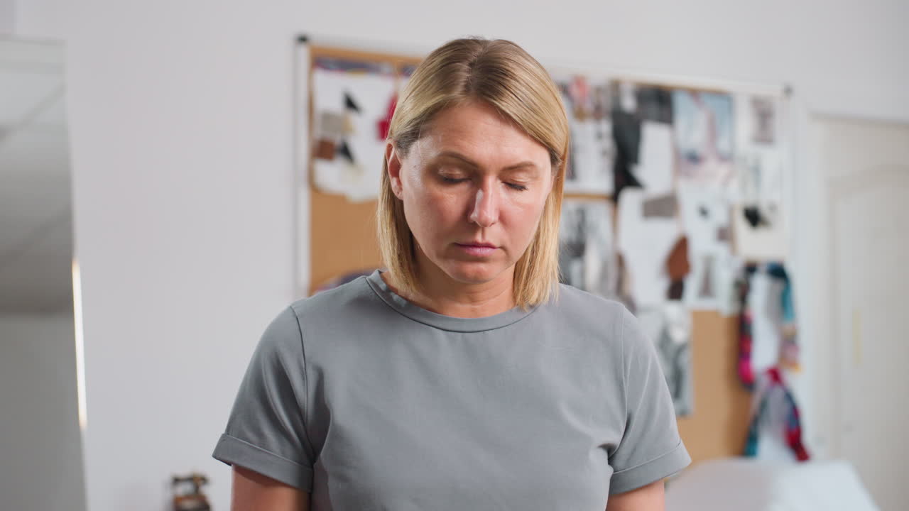Costumer focusing intensely while folding fabric, lifting head up with serious expression in creative workspace, surrounded by blurred background of colorful fabric samples and inspiration boards