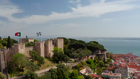 Close-up-aerial-view-of-old-castle-on-the-hill-above-Lisbon-city-center-with-reveal-of-the-sea-with-boats-off-the-Portugal-coast