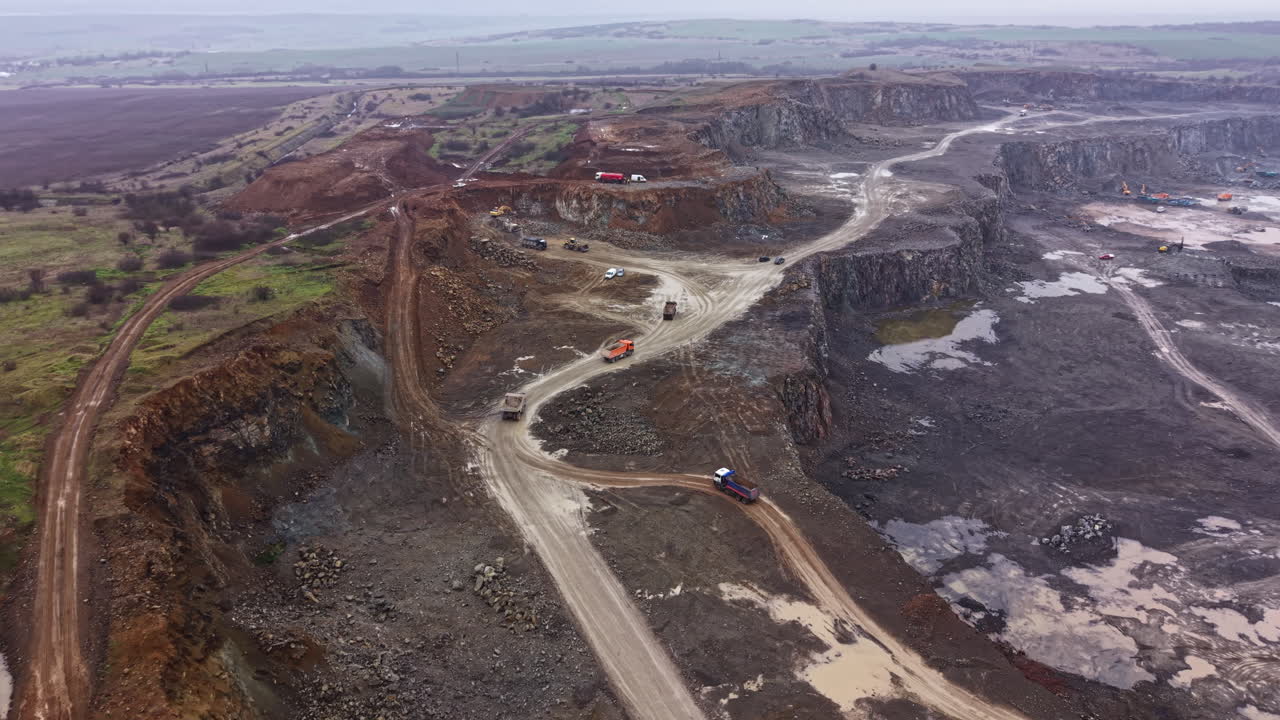 Workers extract granite from a large quarry in a rural area while trucks move materials across dirt roads during cloudy weather