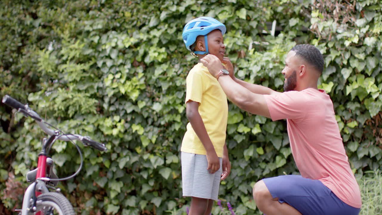 feliz padre afroamericano poniéndose el casco de bicicleta hijo en el jardín, copia espacio, cámara lenta