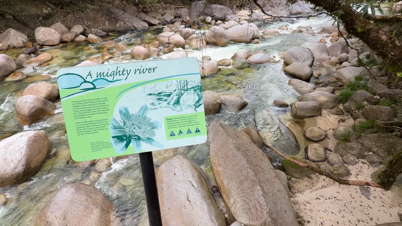 A sign provides information beside a clear, flowing river surrounded by lush greenery and rocks in a rainforest setting