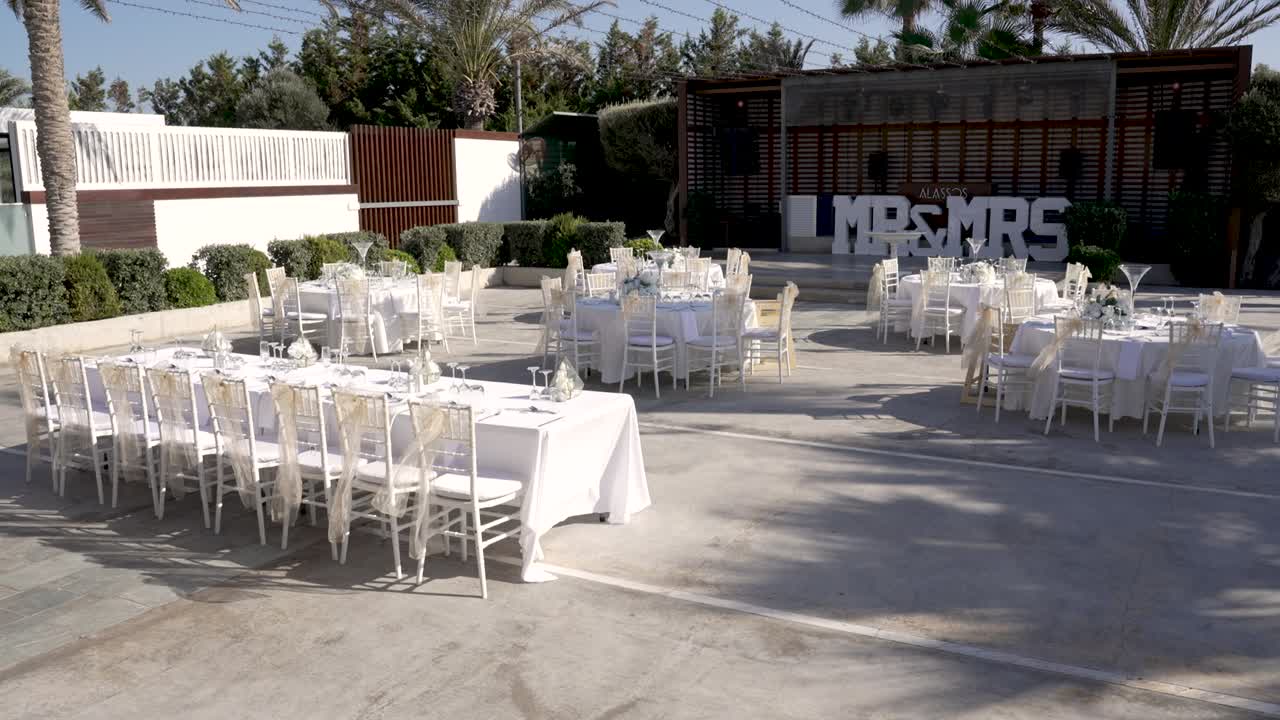 Tables arranged for an outdoor wedding event. Decorations are in place, and the setting has plants and a backdrop with 'Mrs' and 'Mrs'. Time is early afternoon