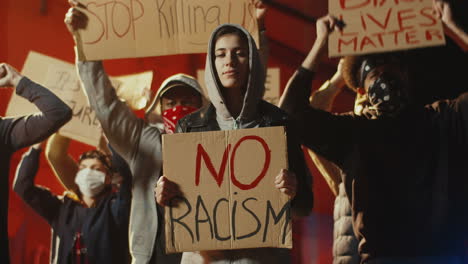 retrato de una mujer caucásica mirando la cámara y sosteniendo un cartel de "no racismo" en una protesta con un grupo multiétnico de personas en la calle"